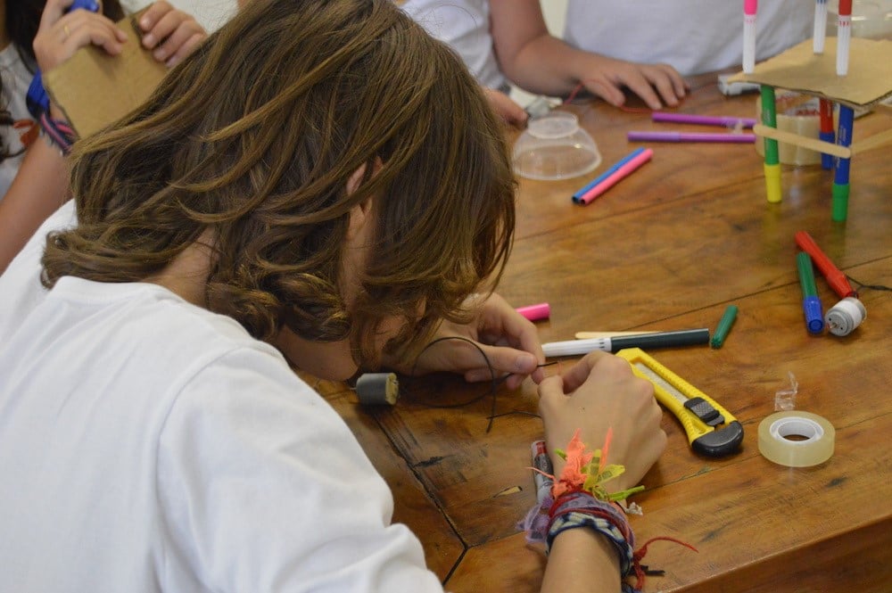 Estudante manuseando um cabo preto sobre uma mesa. Ao redor, estilete, canetas coloridas, durex., demonstrando a importância da cultura maker para a educação.