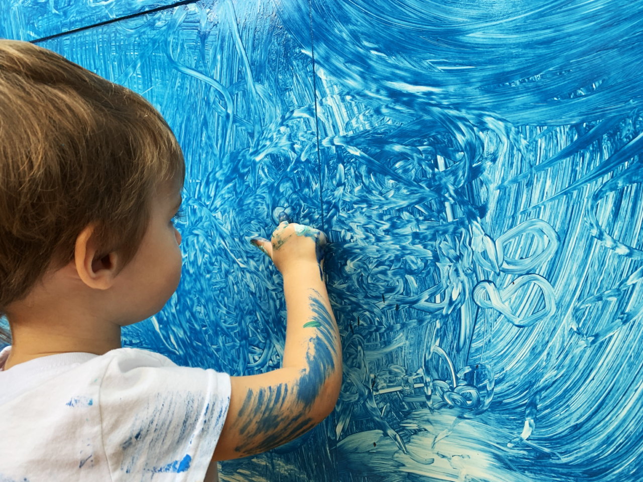 Criança pintando com dedo com tinta azul em uma parede já pintada de azul.