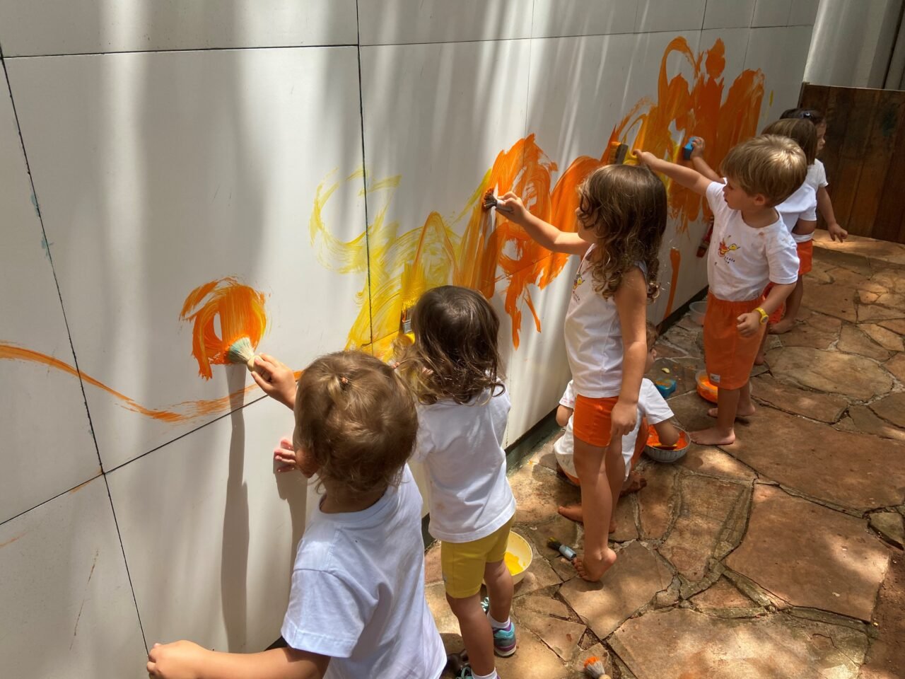 Crianças pequenas pintando a parede com as cores amarela e laranja.